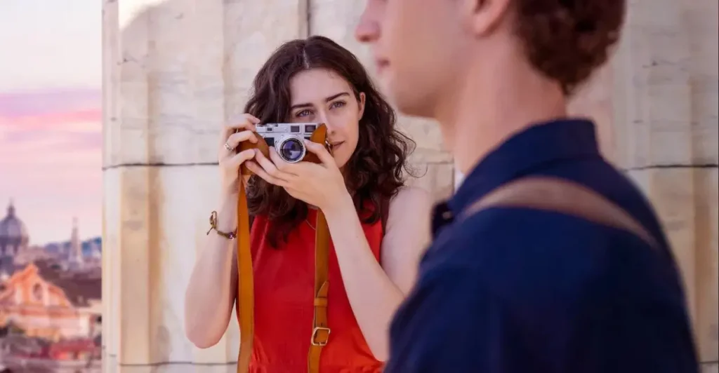 Cena do filme Amor & Gelato (2022), com jovem sorridente fotografando um rapaz em frente a uma paisagem romântica na Itália — romance leve com toques de viagem e descobertas pessoais.
