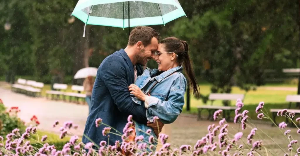 Cena do filme Amor² (2021), com Monika (Adrianna Chlebicka) e Enzo (Mateusz Banasiuk) sorrindo sob a chuva em um parque, protegidos por um guarda-chuva verde — um momento romântico que captura o clima leve e apaixonado da comédia romântica polonesa da Netflix.