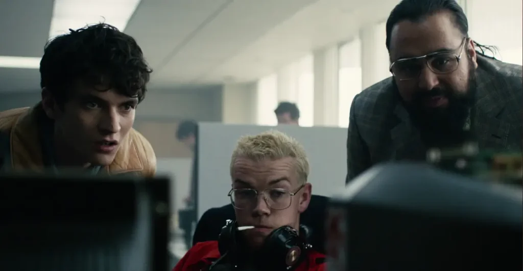 Cena de Black Mirror: Bandersnatch (2018), com os personagens Stefan Butler (Fionn Whitehead), Colin Ritman (Will Poulter) e Mohan Thakur (Asim Chaudhry) observando atentamente a tela de um computador, em momento decisivo do episódio interativo da Netflix que mistura ficção científica, livre-arbítrio e distorção da realidade.
