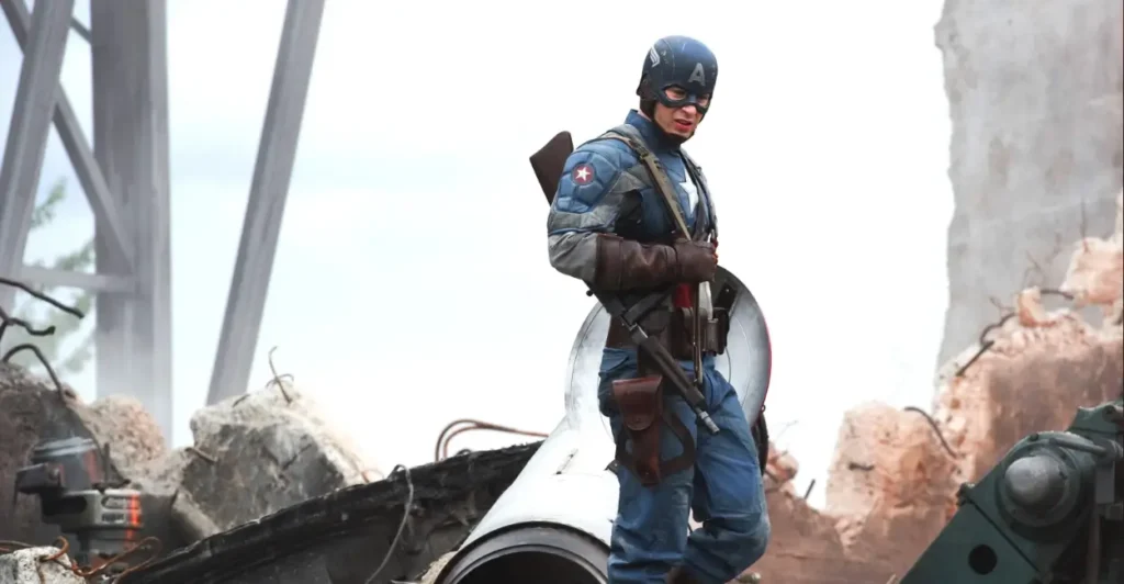 Cena do filme Capitão América: O Primeiro Vingador (2011), com Steve Rogers, o Capitão América (interpretado por Chris Evans), usando seu uniforme clássico da Segunda Guerra Mundial e empunhando seu escudo em meio aos escombros — momento que marca sua luta contra a Hydra no início do Universo Cinematográfico da Marvel.