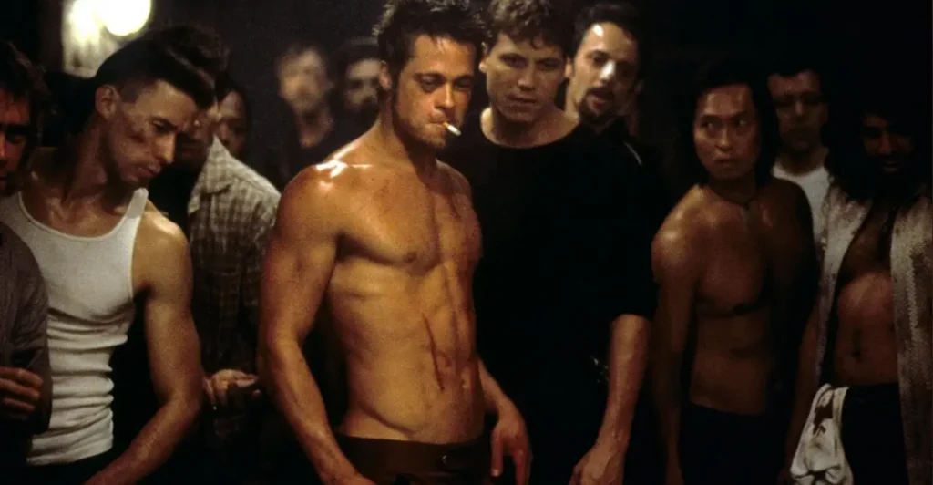 Cena marcante de Clube da Luta (1999), com o personagem Tyler Durden (interpretado por Brad Pitt) no centro de um clube de lutas clandestino, cercado por outros homens ensanguentados.