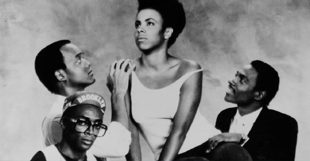 Cena do filme Ela Quer Tudo (1986), com Nola Darling (Tracy Camilla Johns) ao centro, cercada por seus três pretendentes: Jamie Overstreet (Tommy Redmond Hicks), Greer Childs (John Canada Terrell) e Mars Blackmon (Spike Lee), destacando o triângulo amoroso e a independência da protagonista na comédia de Spike Lee.