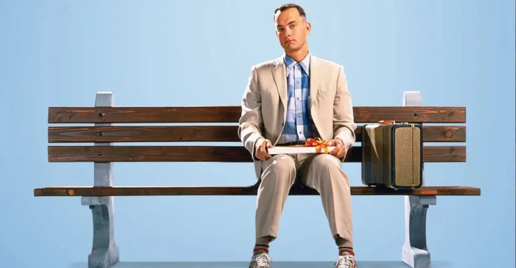Forrest Gump sentado em um banco com uma caixa de chocolates no colo e uma mala ao lado, em cena icônica do filme Forrest Gump (1994), interpretado por Tom Hanks.