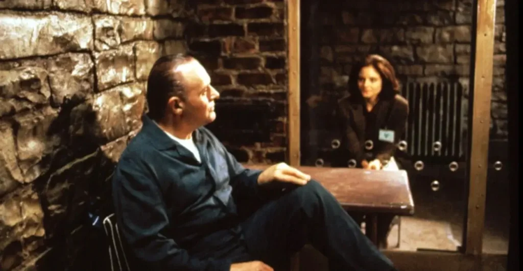 Cena icônica do filme O Silêncio dos Inocentes (1991), com Hannibal Lecter (Anthony Hopkins) sentado em sua cela durante conversa tensa com a agente Clarice Starling (Jodie Foster), separada por uma divisória de vidro — um dos momentos mais memoráveis do suspense psicológico vencedor do Oscar.