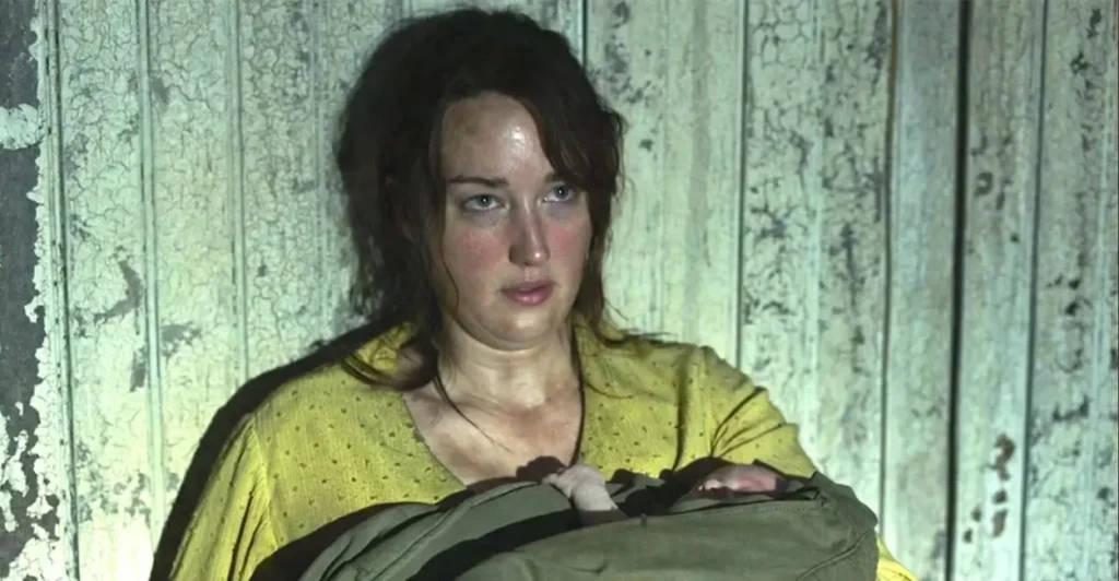Cena da série The Last of Us (2023), com a personagem Anna, interpretada por Ashley Johnson, segurando um bebê recém-nascido em um ambiente deteriorado, simbolizando a luta pela sobrevivência e o vínculo maternal no universo pós-apocalíptico da adaptação da HBO.