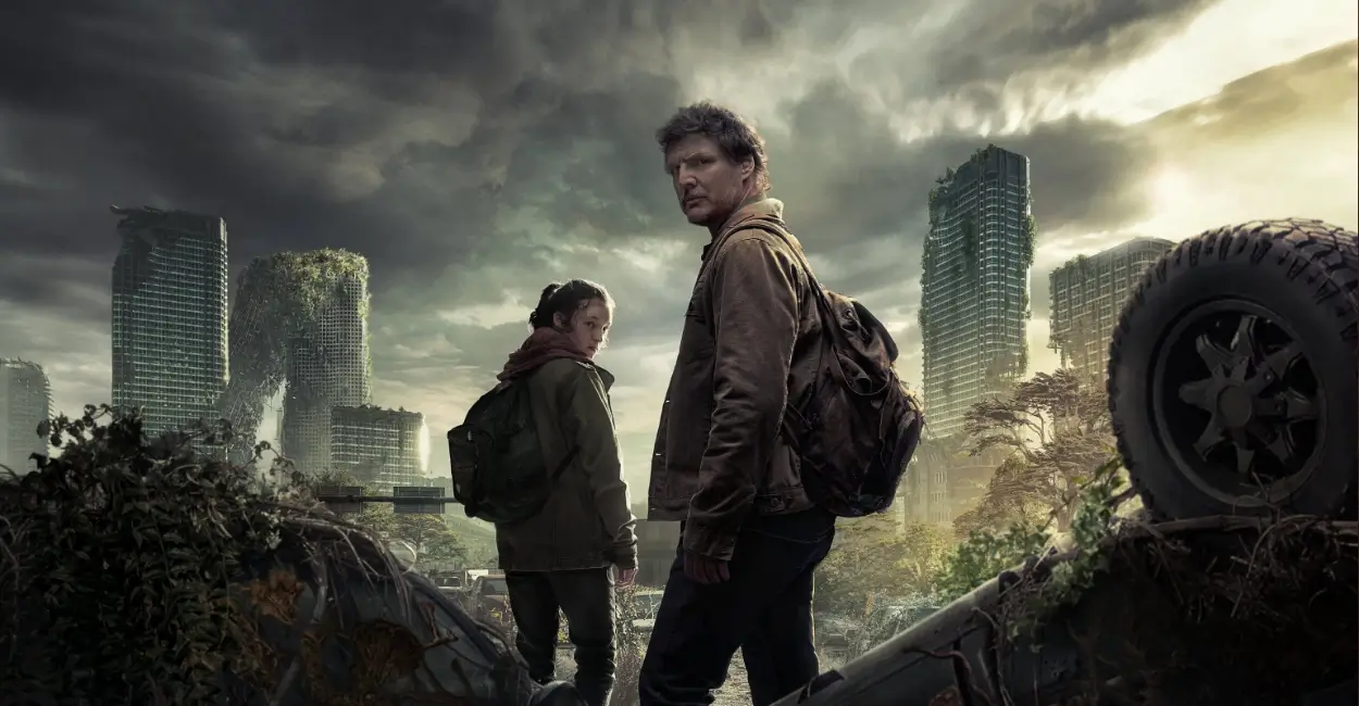 Joel e Ellie caminhando por um mundo pós-apocalíptico, rodeados por prédios destruídos e cobertos de vegetação, em cena da série The Last of Us (2023–atualmente).
