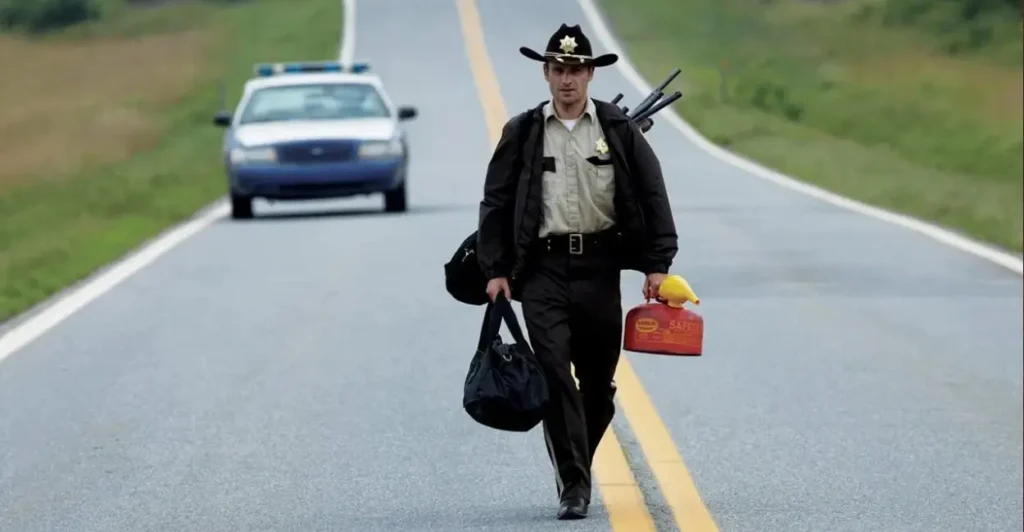 Rick Grimes caminhando por uma estrada deserta em uma cena icônica da série The Walking Dead, vestindo uniforme de xerife e carregando uma lata de combustível.