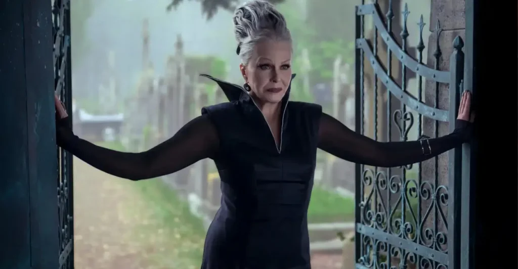 Vovó Addams na série Wandinha da Netflix, com visual sombrio e elegante, abrindo os portões de um cemitério em uma cena misteriosa.