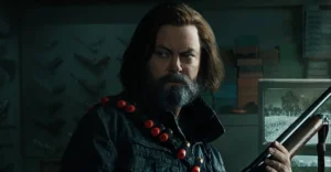 Bill, personagem de The Last of Us, empunha espingarda com cartucheira de munição no ombro, em cena tensa dentro de seu refúgio fortificado.