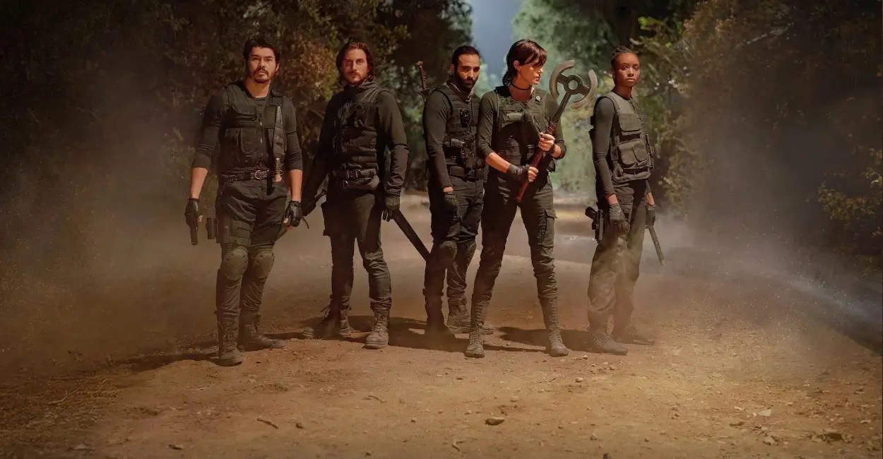 Personagens principais de The Old Guard 2 (2025) prontos para o combate, em uma floresta com visual cinematográfico, vestindo trajes táticos e armados para ação.